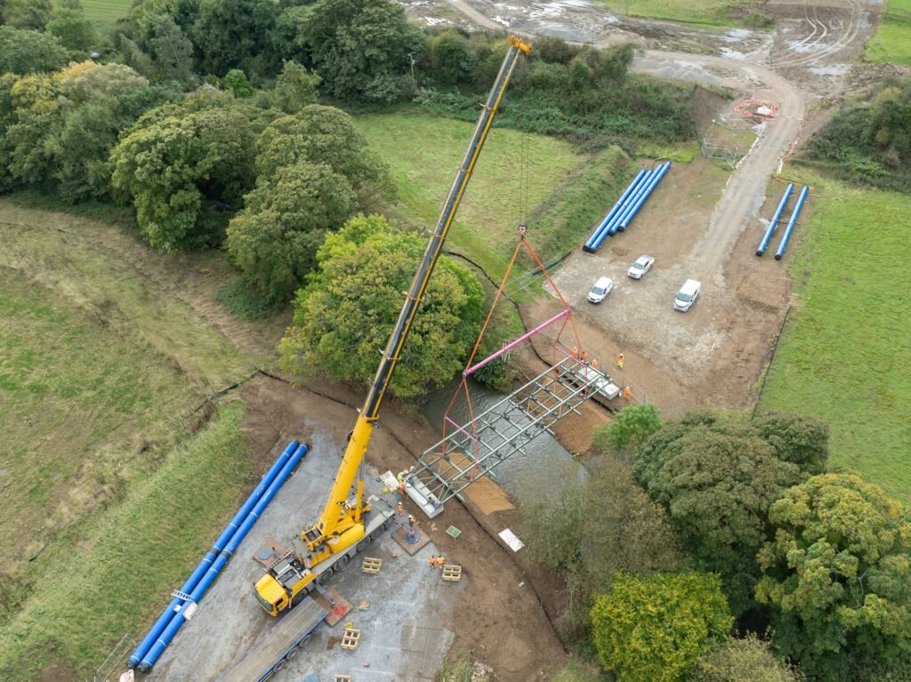 Drone view of a yellow all-terrain crane on a working platform lifting a steel pipe bridge over a wooded beck, with blue pipes laid on the ground