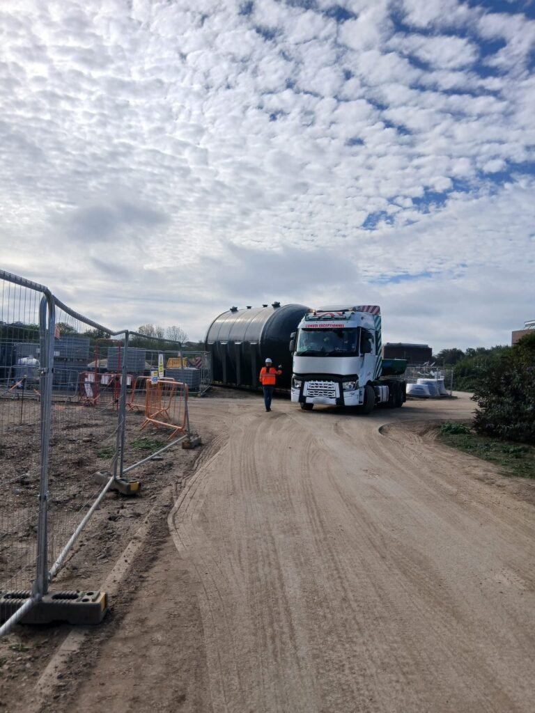 Low loader carrying a sand filter vessel along the site road at Loughborough, guided in by a banksman